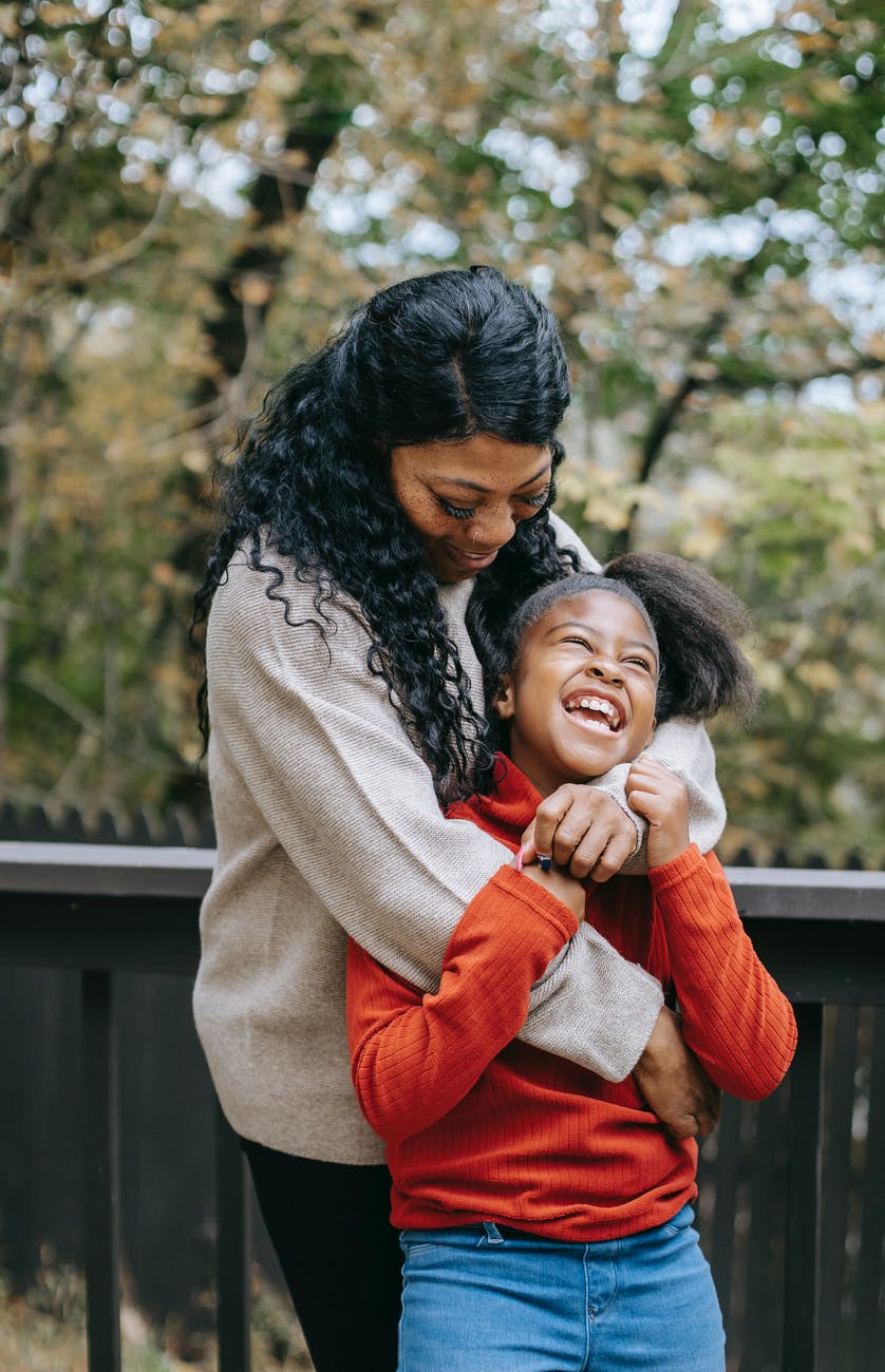 black mother embracing happy daughter near fence